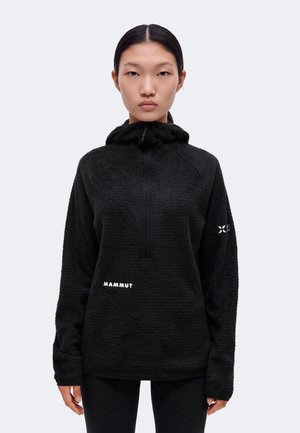 Un mannequin est de face, vêtu d’un sweat à capuche Mammut noir à la texture marquée et d’un pantalon noir, sur un fond clair uni.