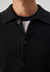 Primer plano de una persona que lleva un cárdigan negro de punto acanalado abotonado sobre una camiseta de rayas blancas y negras.