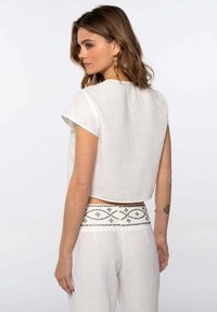 Top corto de lino blanco con mangas cortas, de textura suave y corte holgado. Combinado con pantalones de cintura alta con estampados y detalles metálicos.