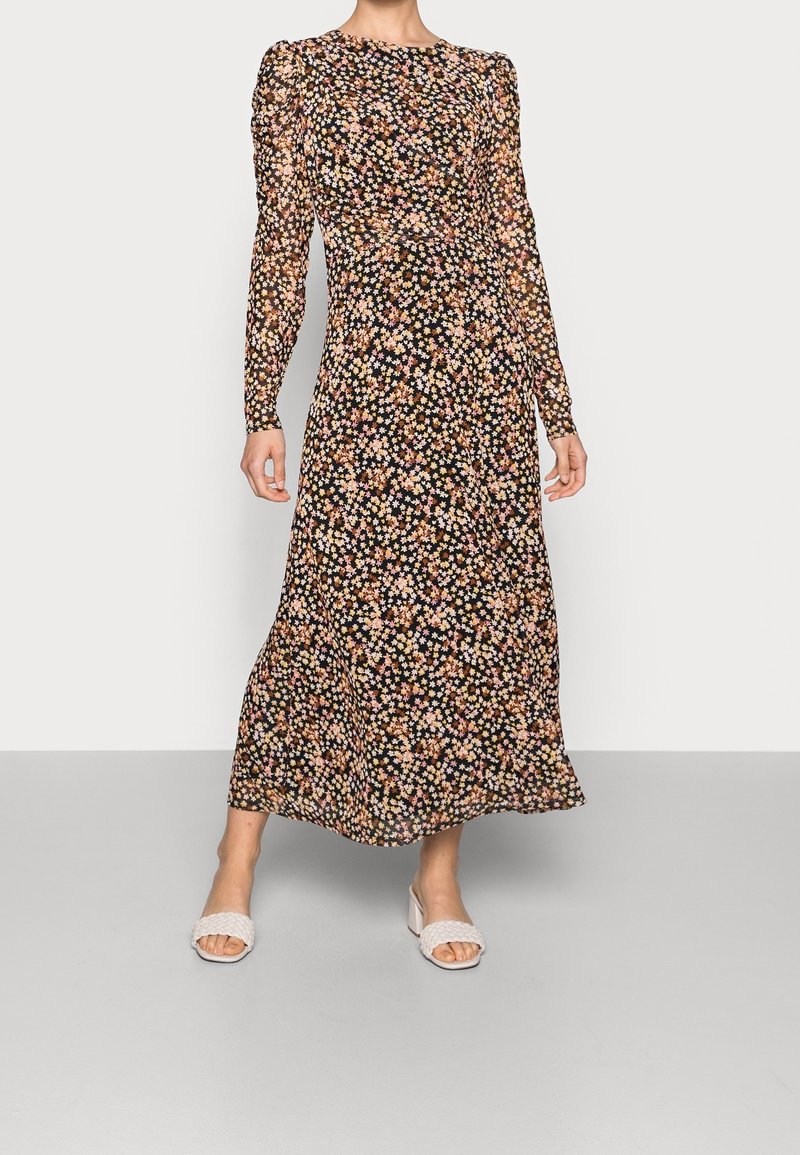 Femme portant une longue robe noire à fleurs avec manches longues et des sandales blanches à talons ouverts, debout sur un sol gris.