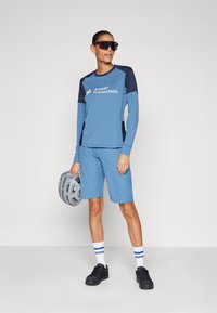 Gai zils garām piedurknēm krekls ar tumši zilām akcentiem, balta logo, apvienots ar gaiši zilu šortiem un melniem tenisa apaviem, turot pelēku ķiveri.