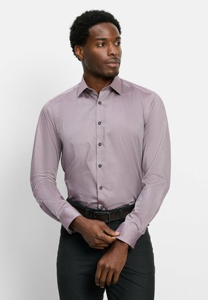 Uomo che indossa una camicia viola a maniche lunghe e pantaloni neri mentre sistema il polsino, guardando di lato su uno sfondo neutro.