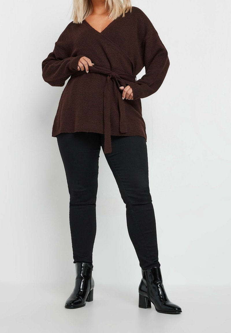 Kobieta w brązowym swetrze z opaską w talii, czarnych obcisłych spodniach oraz czarnych lakierowanych botkach za kostkę stoi na tle białym.