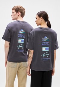 T-shirts de algodão cinza com gola redonda, com um logótipo e texto nas costas em azul e branco. Corte largo com mangas curtas.