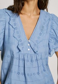 Blusa de algodón de color azul claro con escote en V, bordes de ondas, patrones florales bordados y tres botones decorativos en la parte frontal.