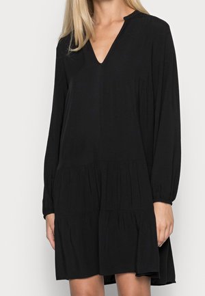 Žena v lehkých černých dlouhých šatech s výstřihem do V a sukni s volány, stojící před jednoduchým světle zbarveným pozadím.