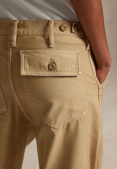 Close-up of person wearing beige pants with buttoned back pocket and hand inside side pocket, showing fabric texture and stitching details.