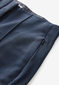 Marineblauwe broek gemaakt van gestructureerde stof, met een zijzak met een zwarte ritssluiting. Het ontwerp bevat voorplooien en strakke lijnen.