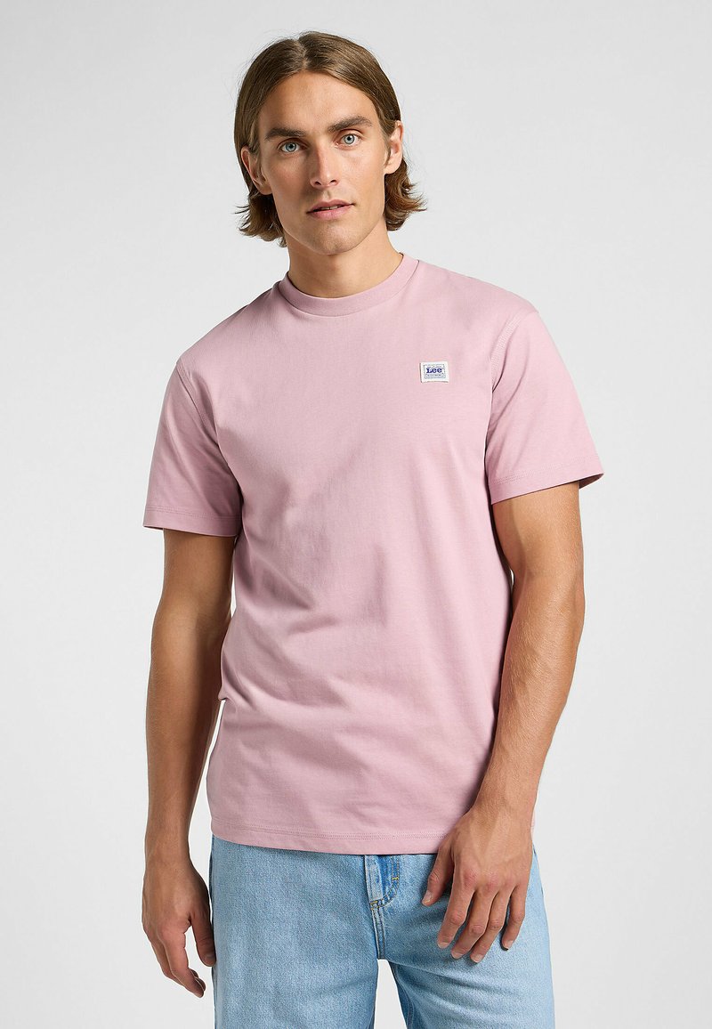 Lyserød bomulds t-shirt med rund hals, korte ærmer og et lille blåt og hvidt logo-patch på brystet. Sammen med blå denim jeans.