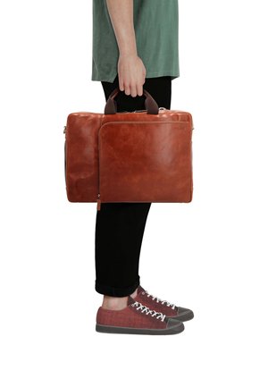 Person, die eine braune Lederaktentasche an den Griffen hält, trägt schwarze Hose, grünes Hemd und rote Turnschuhe.