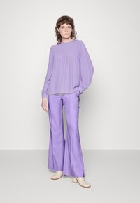 Blusa lavanda com decote franzido e mangas largas, combinada com calças lavanda retas. Os sapatos são claros com um salto bloco.