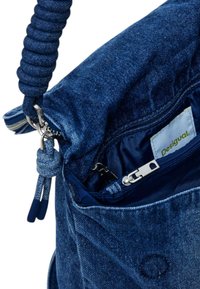 Blaue Jeans-Tasche mit einem strukturierten, gewebten Griff. Das Innere verfügt über eine Reißverschlusstasche und ein Etikett mit "Desigual" auf hellem Hintergrund.