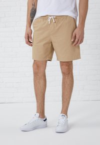 Beige Baumwollshorts mit elastischem Bund und weißem Kordelzug, im Standardfit und mit schlichter Optik, kombiniert mit weißen Sneakers.