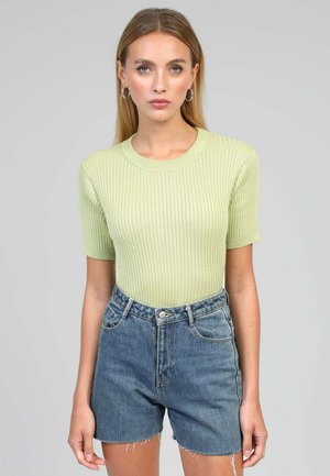 Femme aux longs cheveux blonds portant un haut vert clair côtelé à manches courtes et un short en denim bleu taille haute sur un fond uni.