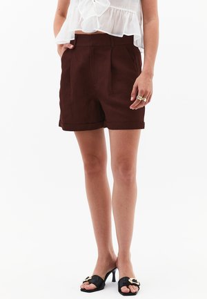 Shorts en lin marron avec des plis, ourlets retroussés et poches latérales, associés à des talons ouverts noirs avec un accent doré.