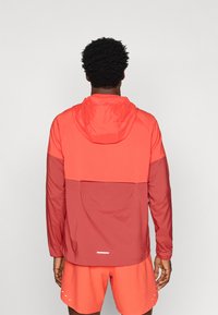 Κόκκινο και κοραλλί ελαφρύ μπουφάν με κουκούλα, χαρακτηρισμένο από χρωματική μπλοκ διάταξη, ανακλαστικές λεπτομέρειες και πίσω τσέπη.