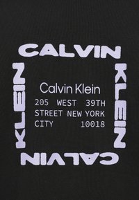 Μαύρο ύφασμα με λευκό τυπωμένο κείμενο. Διακρίνεται έντονα το "CALVIN KLEIN", περιβαλλόμενο από ορθογώ�νιο πλαίσιο με μια διεύθυνση.