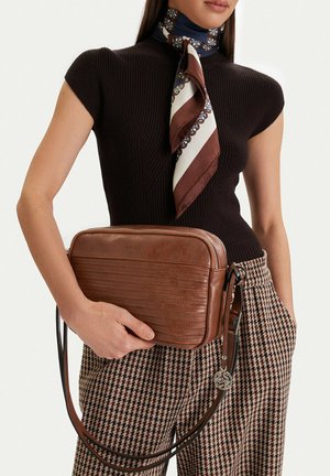 Žena v černém tričku s krátkým rukávem, se šátkem s motivem a kostkovanými kalhotami drží hnědou koženou kabelku přes rameno v úrovni pasu.