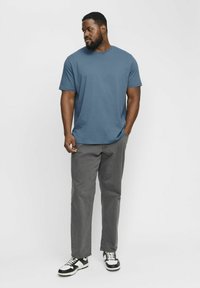Camiseta de algodón azul con cuello redondo, combinada con pantalones grises y zapatillas de deporte en blanco y negro. El modelo está de pie con una mano en el bolsillo.
