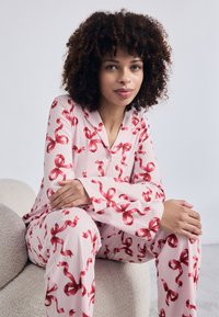 Conjunto de pijama rosa com padrões de fita vermelha, mangas longas e decote abotoado. Tecido suave e ajuste descontraído, estilizado com colarinho redondo.