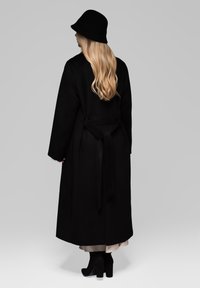 Musta villa takki pitkillä hihoilla ja vyötäröltä sidottuna, yhdistettynä samanlaiseen mustaan ämpärihattuun ja nilkkapituisilla mustilla korkokengillä.