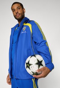 Blauw sportjack met gele accenten, gecombineerd met een blauw overhemd en een blauwe broek. Houdt een zwart-witte voetbal vast met sterpatronen.