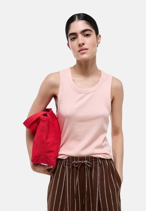 Femme aux cheveux foncés portant un haut rose sans manches et un pantalon marron rayé, tenant un sac rouge sur un fond blanc uni.
