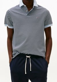 Polo rayé bleu marine et blanc avec un col bleu clair, manches courtes, et un logo sur le côté gauche. Associé à un pantalon de jogging noir.