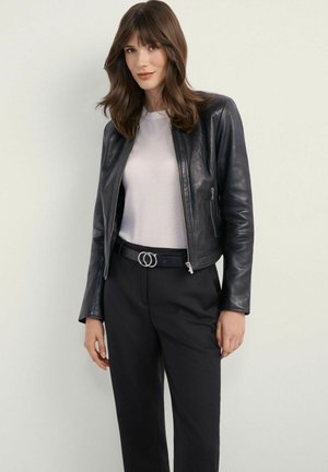 Kobieta o brązowych włosach, ubrana w czarną skórzaną kurtkę na zamek, białą koszulę, czarne spodnie i czarny pasek z podwójną klamrą, stojąca na jasnym tle.