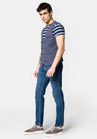 Pasek w niebiesko-białe paski pod szarym kamizelką w kratę; dopasowane dżinsy i szare trampki z białymi podeszwami; w pozycji stojącej.