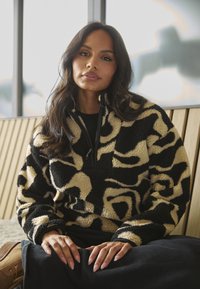 Pull-over en polaire noir et beige avec un col à demi-zip, arborant un motif en tourbillon. La texture est douce et moelleuse. Porté avec un pantalon noir.