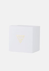 Bijela teksturirana kutija s zlatnim trokutastim logotipom koji sadrži riječ "GUESS" i upitnik. Kvadratnog oblika s čistim dizajnom.