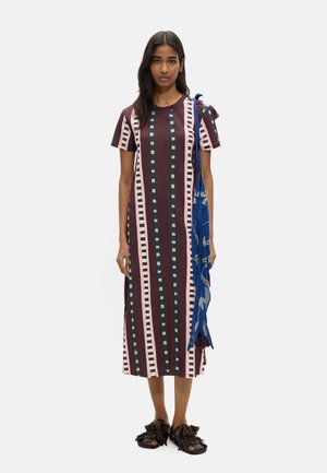 Kobieta stojąca przodem, ubrana w brązową sukienkę w pionowe różowe i zielone paski, czarne sandały z frędzlami oraz niebieską torebkę na ramię.