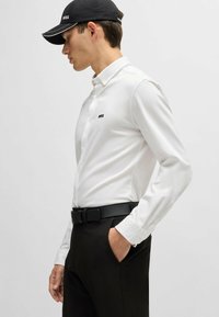 White, long-sleeve shirt with a black logo on the chest, paired with black trousers and a black cap, featuring a clean, tailored design.