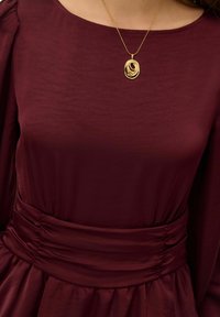 Vijolična obleka iz gladkega blaga z nabranim pasom; vsebuje zlato ogrlico z obeskom z več slojnimi ovalnimi oblikami.