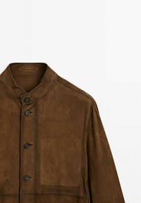 Veste en daim marron avec un col montant, dotée de boutons noirs et d'une poche poitrine. Texture lisse avec un design minimaliste.