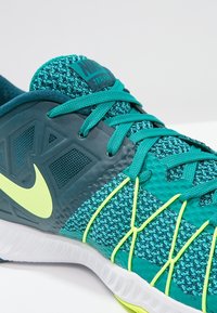 Una zapatilla deportiva en color azul verdoso con una parte superior de malla transpirable, paneles texturizados, un prominente swoosh amarillo de Nike y cordones vibrantes de contraste.