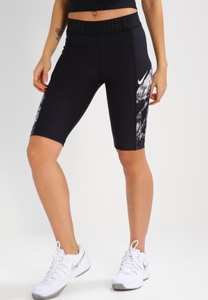 Femme portant un cuissard de cyclisme noir Nike avec logo blanc et motif sur les côtés, associé à des chaussures de sport blanches Nike sur un fond uni.