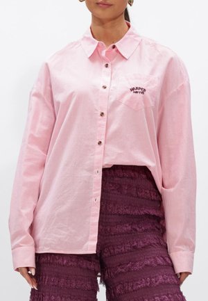 Vrouw draagt een lichtroze overhemd met knopen en "HARPER AND YVE" op het borstzakje en paarse kanten gelaagde shorts.