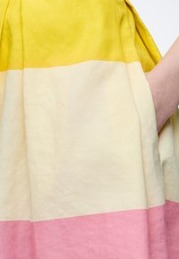 Χέρι που ακουμπάει μέσα στην τσέπη μιας τρίχρωμης φούστας με οριζόντιες ρίγες σε κίτρινο, κρεμ και ροζ.