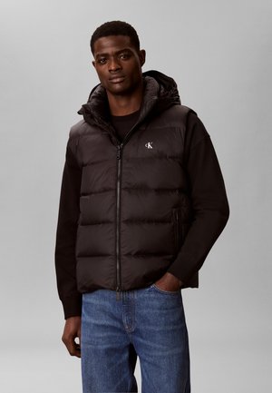 Mand iført sort dunvest med hætte over sort langærmet skjorte og blå jeans, stående med den ene hånd i lommen mod en grå baggrund.