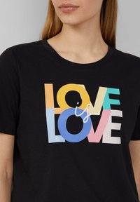 T-shirt noir à manches courtes avec un design en texte multicolore qui indique "L'AMOUR est L'AMOUR" dans des teintes vibrantes de jaune, bleu et rose.