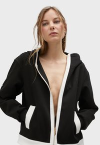 Crna zip-up dukserica s dubokim prednjim otvaranjem, bijelim akcentima na manžetama i džepovima, s mekanom teksturom i kapuljačom. Zlataasti detalj na zipu.