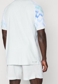 Takav kuva peronje osoby svieti biele sportovne oblečenie s modrými geometrickými vzormi na rukávoch a textom "DRI-FIT ADV" blízko spodného okraja.