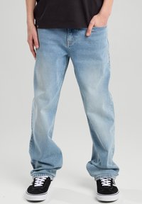 Light blue denim jeans with a relaxed fit, single front pocket, and frayed hem. Paired with black sneakers featuring white laces.
