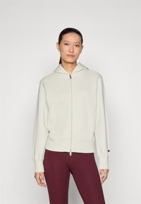 Light beige zip-up hoodie with a hood, ribbed cuffs and hem, and a small logo on the chest. Paired with dark maroon leggings.
