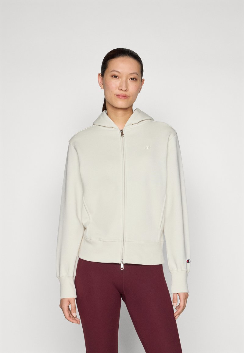 Light beige zip-up hoodie with a hood, ribbed cuffs and hem, and a small logo on the chest. Paired with dark maroon leggings.