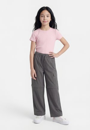 Tüdruk, kellel on pikad mustad juuksed, seljas roosa soonikkoeline lühikese varrukaga topp, hallid cargo-püksid ja valged tossud, seisab ühe käega puusal.