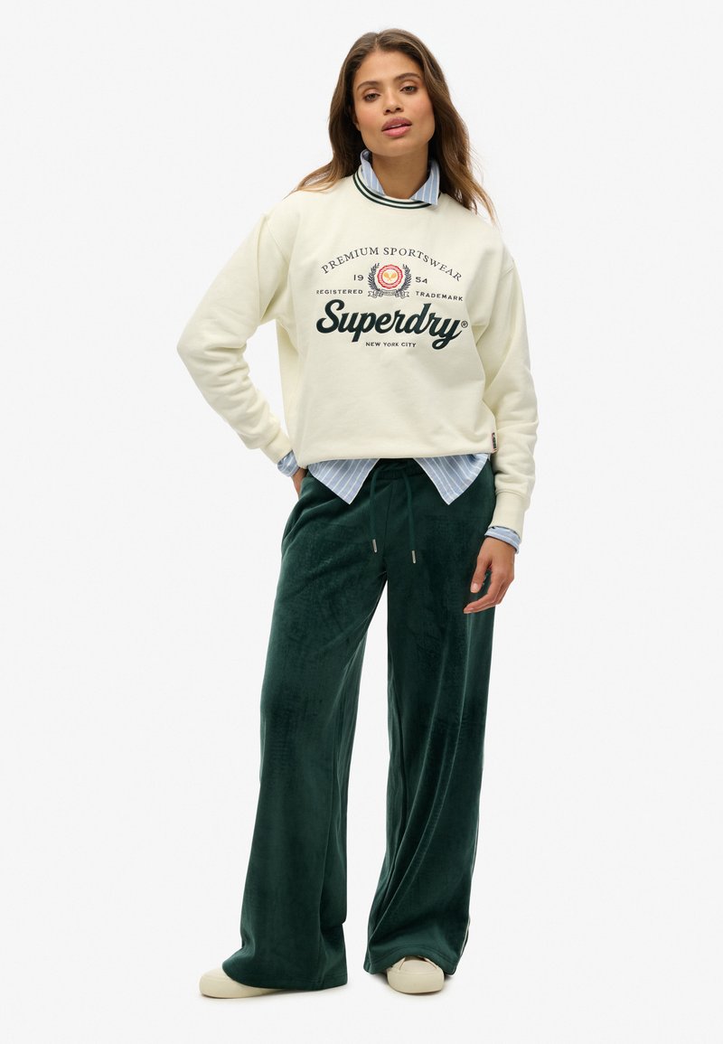 Creme sweatshirt med "Superdry" logo, parret med grønne velour bredde bukser. Lyseblå stribet skjorte underneden. Bløde teksturer.