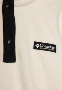 Kremfarget fleecegenser med svart trykknappslukking og krage. Har en vevd Columbia Sportswear Company-merkelapp foran. Myk tekstur.